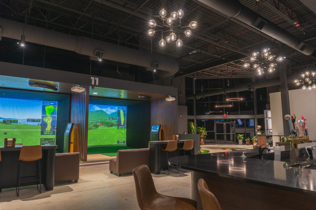 Interior view of the Play Golf VX venue, showcasing a sophisticated indoor golf simulator setup. The image features spacious playing areas with advanced technology screens, elegantly designed seating arrangements for relaxation and socializing, and a vibrant atmosphere with ambient lighting.